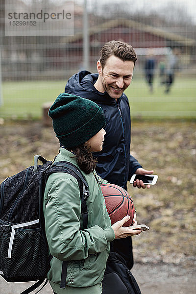 Lächelnder Vater und Sohn unterhalten sich beim Gehen auf der Straße nach dem Basketballtraining im Winter