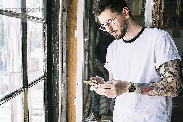 Selbstbewusster männlicher Hipster-Hacker-Textnachrichten per Smartphone durch Fenster im Kreativbüro