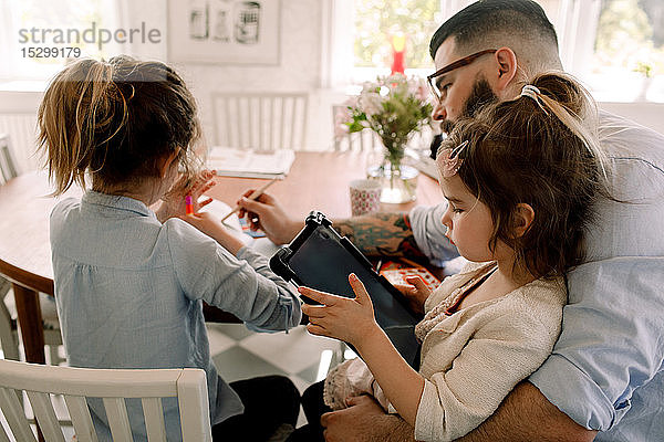Mädchen benutzt digitales Tablett  während der Vater der Tochter bei den Hausaufgaben am Esstisch hilft