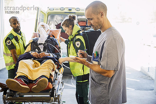 Männliche Krankenschwester schreibt Bericht  während Sanitäter den Patienten im Krankenhaus auf die Trage schieben