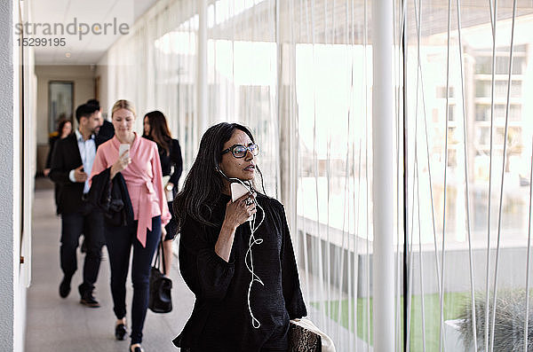 Geschäftsfrau spricht mit einem Smartphone auf dem Flur  während sie das Büro mit Kollegen im Hintergrund verlässt