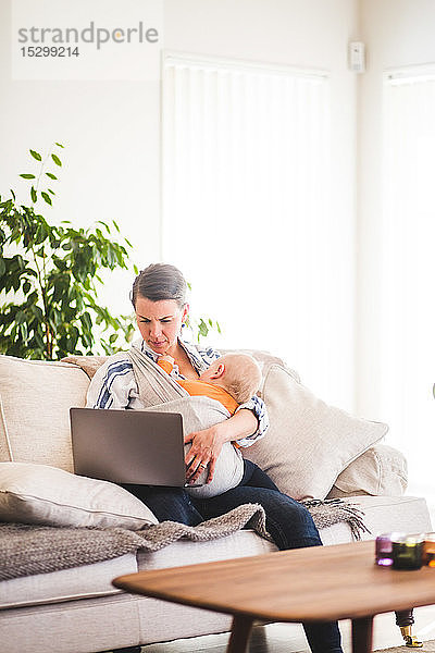 Multi-Tasking-Mutter benutzt Laptop  während sie ihr Baby auf dem Sofa im Heimbüro betreut