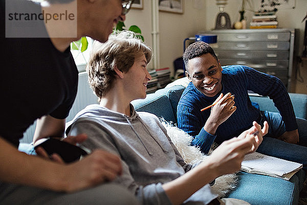 Lächelnder Teenager zeigt Freunden sein Handy auf dem Sofa  während er zu Hause lernt