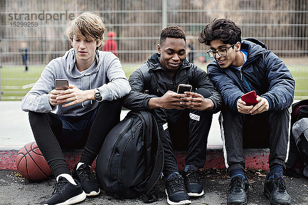 Social-Media-süchtige Freunde benutzen Mobiltelefone  während sie nach dem Basketballtraining in der Stadt auf dem Bürgersteig sitzen