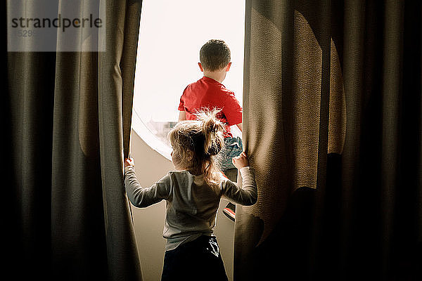 Rückansicht eines Mädchens  das einen Vorhang hält  während der Bruder am Fenster im Hotelzimmer sitzt