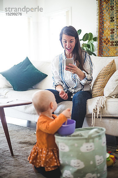 Glückliche berufstätige Mutter fotografiert spielende Tochter während der Telearbeit im Wohnzimmer