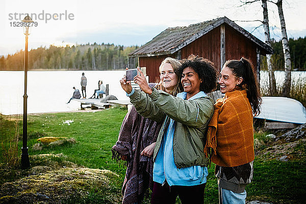 Glückliche Freundinnen  die sich mit einem Smartphone selbstständig machen  während sie bei Sonnenuntergang auf einem Grasfeld am See stehen