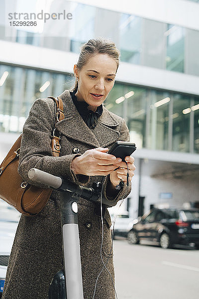 Selbstbewusste Geschäftsfrau benutzt ein Smartphone  während sie mit einem elektrischen Roller in der Stadt steht