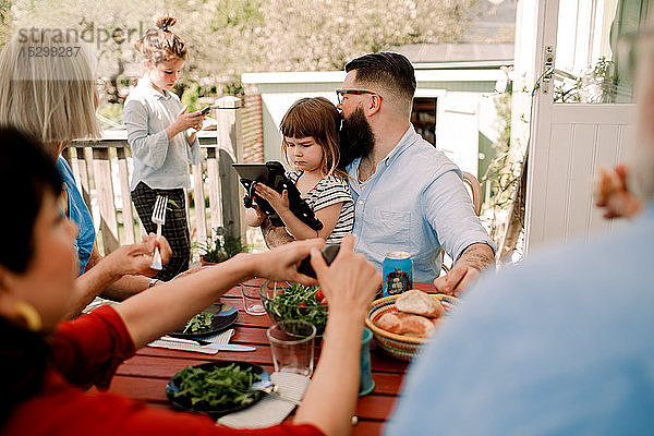 Mehrgenerationen-Familie  die Nahrung zu sich nimmt  während die Töchter Technologie auf der Terrasse nutzen