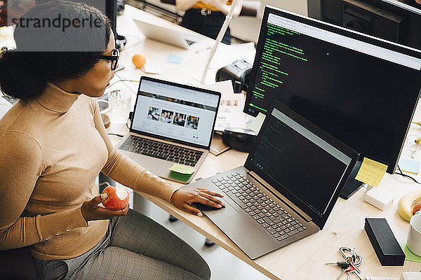 Hochwinkelansicht einer IT-Fachfrau  die einen Apfel hält  während sie einen Laptop auf dem Schreibtisch im Kreativbüro benutzt