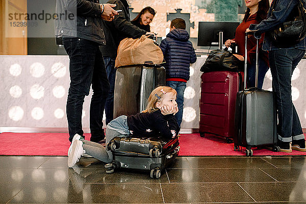 Langweiliges Mädchen schaut weg  während es sich an einen Koffer lehnt  während die Familie an der Rezeption im Hotel steht