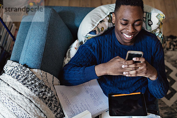 Schrägansicht eines sozialmediensüchtigen Teenagers  der ein Smartphone benutzt  während er auf dem Sofa mit Büchern im Wohnzimmer liegt