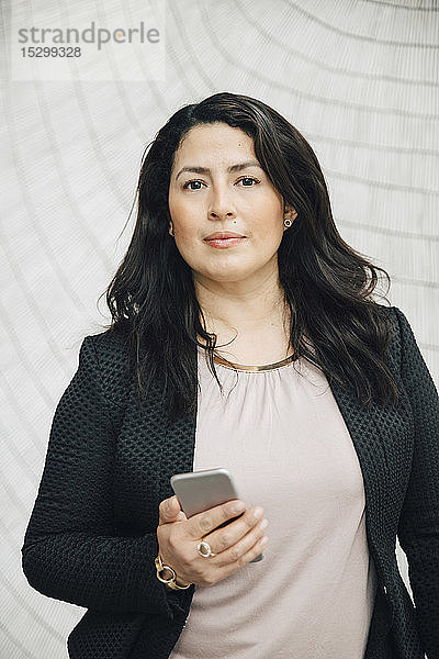 Porträt einer selbstbewussten Geschäftsfrau mit einem Smartphone in der Hand  die im Büro an der Wand steht