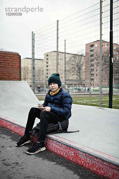 Ganzfigur eines Jungen in warmer Kleidung auf einem Rucksack sitzend in voller Länge vor einem Fussballfeld in der Stadt