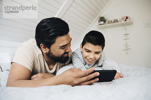 Vater und lächelnder behinderter Sohn sehen sich zu Hause im Bett liegend über Handy einen Film an