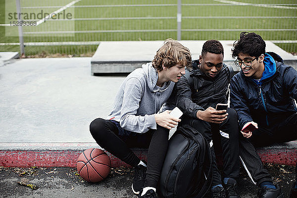 Männliche Freunde benutzen Mobiltelefone  während sie nach dem Basketballtraining im Winter auf dem Bürgersteig sitzen