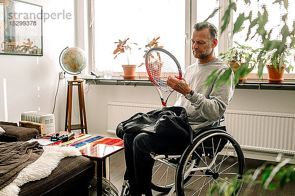 Nachdenklicher  reifer Mann hält Tennisschläger  während er zu Hause im Rollstuhl sitzt