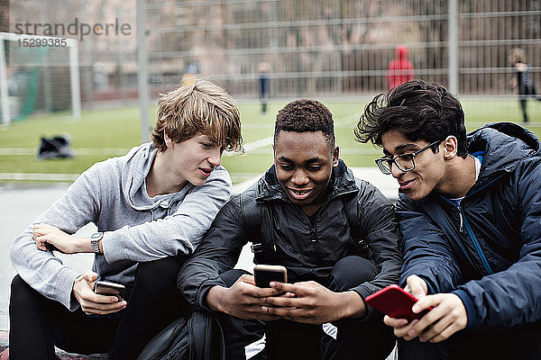 Lächelnde Freunde  die auf das Telefon eines Teenagers schauen  während sie am Fußballfeld sitzen