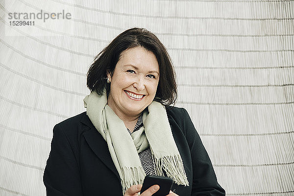 Porträt einer lächelnden  reifen Geschäftsfrau mit einem Smartphone in der Hand  die im Büro an der Wand steht