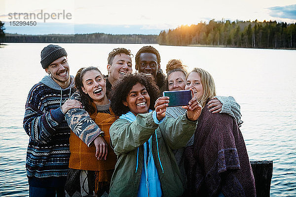 Glückliche männliche und weibliche Freunde  die bei Sonnenuntergang am See stehen und sich mit einem Smartphone selbstständig machen