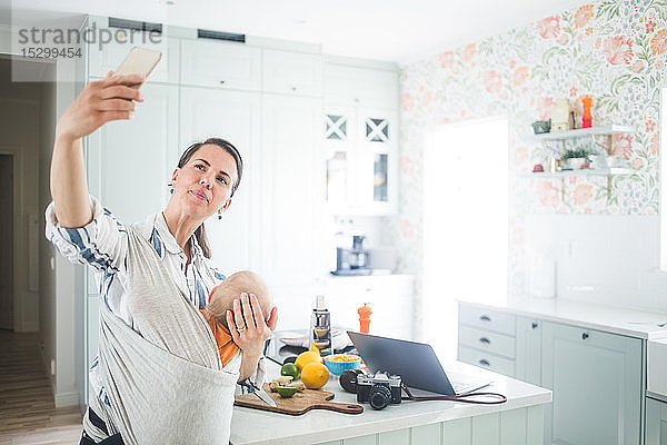 Weibliche Freiberuflerin bloggt über Smartphone  während sie ihre Tochter in der Küche zu Hause trägt