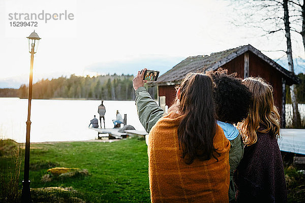 Freundinnen  die sich mit einem Smartphone selbstständig machen  während sie bei Sonnenuntergang auf einem Grasfeld am See stehen