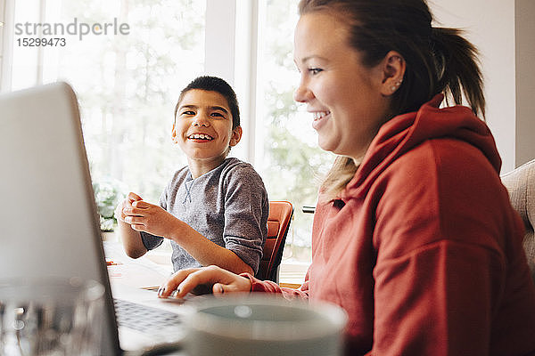 Mutter benutzt Laptop  während sie mit ihrem glücklichen autistischen Sohn zu Hause im Wohnzimmer sitzt