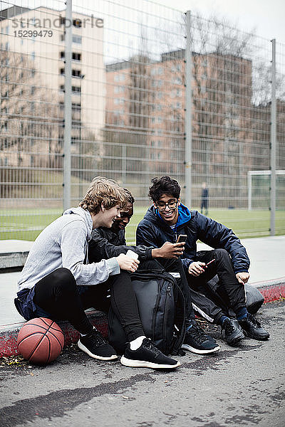 Social-Media-süchtige Freunde in voller Länge  die warme Kleidung tragen  während sie nach dem Basketballtraining auf dem Bürgersteig sitzen d