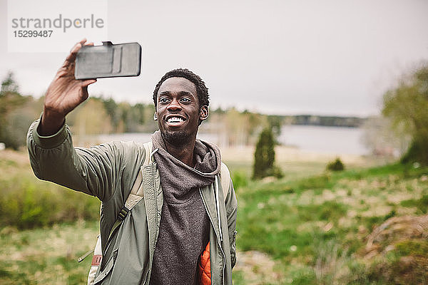 Lächelnder junger Mann  der sich mit einem Smartphone selbstständig macht  während er auf einer Landschaft gegen den Himmel steht