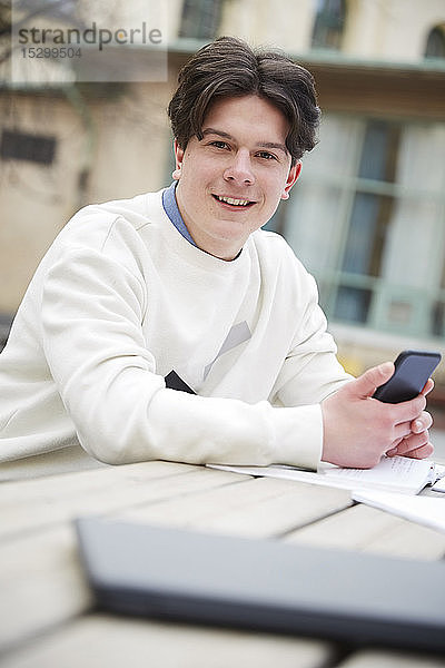 Porträt eines selbstbewussten Teenagers mit Handy in der Hand  der am Tisch auf dem Schulhof sitzt