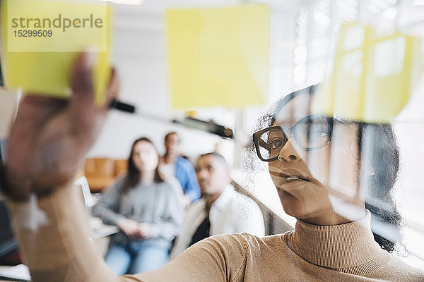 Computerhackerin klebt Haftnotizen auf Glas  während ihre Kollegen im Hintergrund sitzen