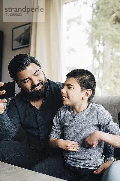 Lächelnder Vater hält Handy in der Hand und schaut den autistischen Sohn an  während er auf dem Sofa im Wohnzimmer sitzt