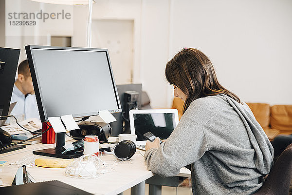 Seitenansicht eines weiblichen Programmierers  der im Büro sitzend ein Mobiltelefon am Schreibtisch benutzt