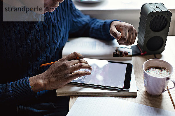 Mittelsektion eines Teenagers  der bei den Hausaufgaben ein digitales Tablett mit einer Kaffeetasse auf dem Tisch benutzt