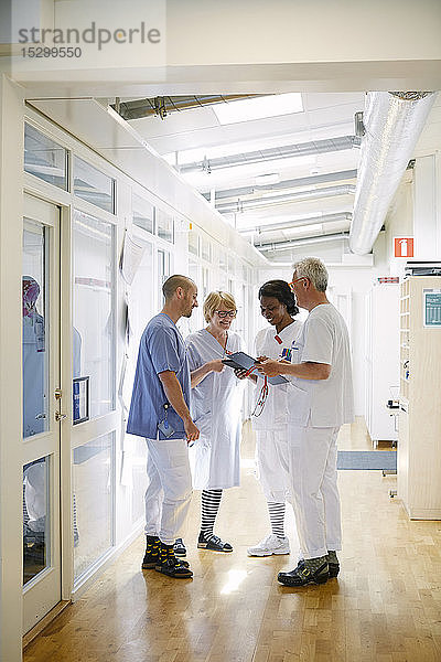Mediziner diskutieren im Krankenhauskorridor stehend über ein digitales Tablett in voller Länge