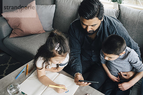 Hochwinkelaufnahme eines Vaters  der seinem Sohn ein Mobiltelefon zeigt  während die Tochter zu Hause in einem Buch zeichnet