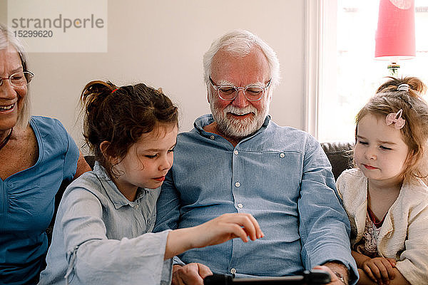 Lächelnde Großeltern sitzen mit Enkelkindern  während sie zu Hause im Wohnzimmer auf ein digitales Tablet schauen