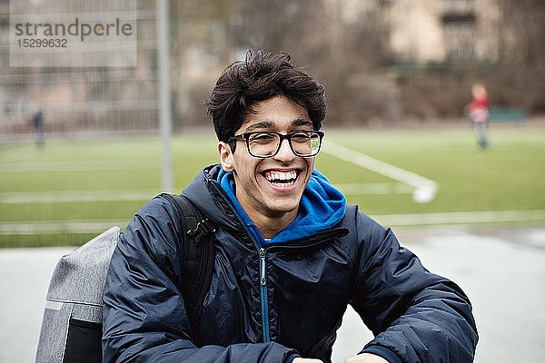 Fröhlicher junger Mann in warmer Kleidung und mit Brille am Fußballfeld in der Stadt sitzend