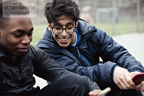 Lächelnder junger Mann  der das Handy eines Freundes gegen ein Spielfeld in der Stadt im Winter betrachtet
