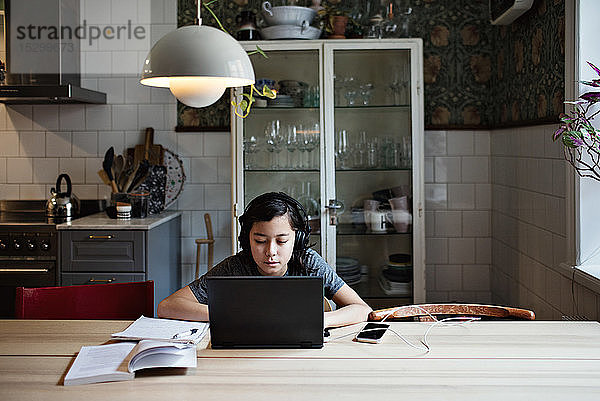 Ernsthafter Junge trägt Kopfhörer  während er bei den Hausaufgaben zu Hause einen Laptop benutzt