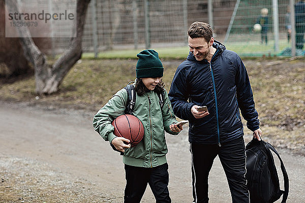 Glückliche Vater und Sohn benutzen Mobiltelefone  während sie nach dem Basketballtraining im Winter auf der Straße gehen