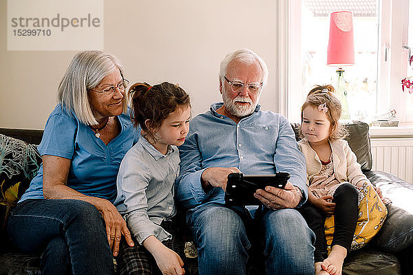 Großeltern sitzen mit Enkelkindern zusammen  während sie zu Hause im Wohnzimmer ein digitales Tablet benutzen