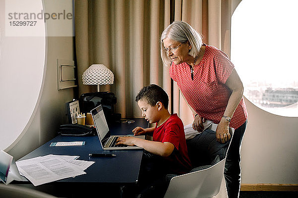 Großmutter sieht ihren Enkel am Laptop an  während sie im Hotelzimmer am Tisch sitzt