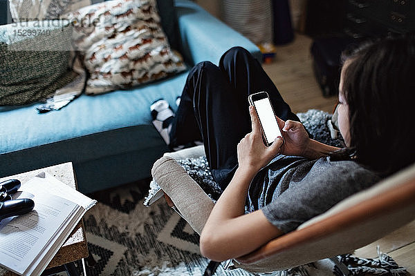 Schrägansicht eines Jungen  der ein Mobiltelefon benutzt  während er im Wohnzimmer sitzt