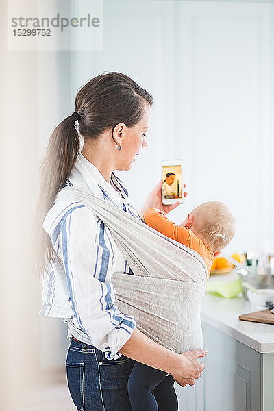 Seitenansicht einer weiblichen Bloggerin  die Fotobotschaften verschickt  während sie ihre Tochter zu Hause in der Küche trägt