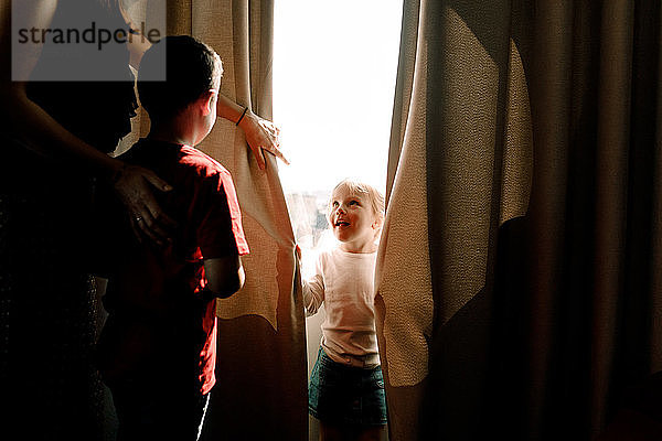 Mädchen steht am Fenster und schaut Mutter mit Junge im Hotelzimmer an