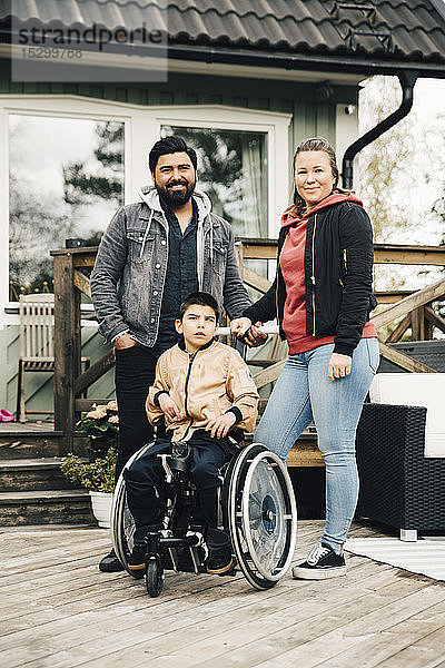 Porträt von lächelnden Eltern  die neben einem behinderten Sohn stehen  der im Rollstuhl gegen das Haus sitzt
