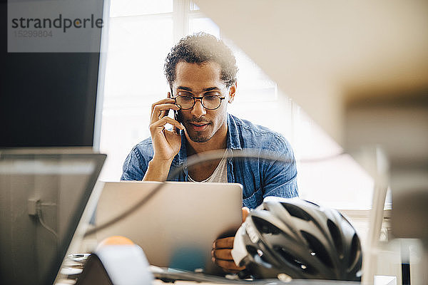 Computerprogrammierer  der sich mit einem Smartphone unter Verwendung eines Laptops im Kreativbüro unterhält