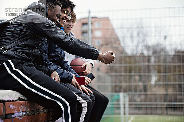 Glückliche männliche Freunde  die nach dem Basketballtraining im Winter auf der Stützmauer sitzen