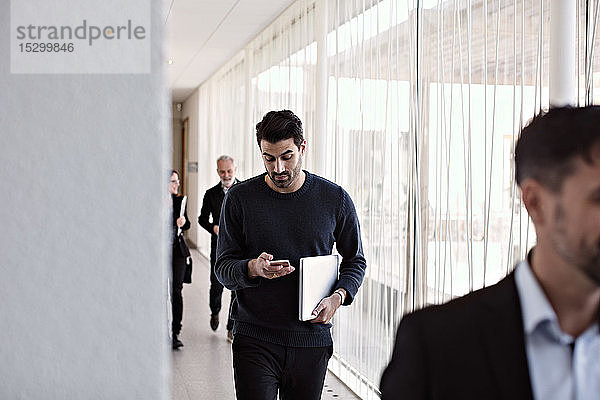 Geschäftsmann benutzt ein Smartphone  während er beim Verlassen des Büros mit Kollegen im Korridor geht